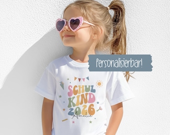 Puede incluir: Una camiseta blanca con el texto "SCHUL KIND 2026 Amelie" en letras y gráficos coloridos. La camiseta presenta un sol, estrellas y un diseño de banner. La niña lleva gafas de sol en forma de corazón.