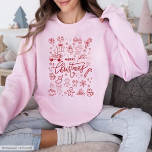 Könnte beinhalten: Ein hellrosa Langarm-Sweatshirt mit einem roten Grafikdruck auf der Vorderseite. Das Design enthält den Text "MERRY Christmas", umgeben von verschiedenen weihnachtlichen Symbolen wie Schneeflocken, einem Lebkuchenmann, einem LKW und Geschenken.