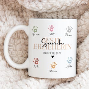 May include: White ceramic mug with a large handle, featuring colorful handprints and the text "Beste Erzieherin Sarah". Additional text reads "Danke f&uuml;r die tolle Kita Zeit" with a small black heart. Names are written below the handprints.