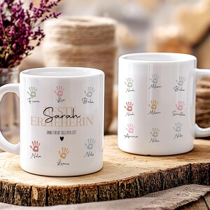 May include: Two white ceramic mugs decorated with colourful handprints. One mug features the name "Sarah" and the text "BESTE ERZIEHERIN" with other names and handprints. The other mug has multiple names and handprints. The mugs are on a wooden surface.