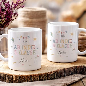 May include: Two white ceramic mugs with the text "AB IN DIE 5. KLASSE" in pastel colours, the year 2026 and the name "Nieke". The mugs are decorated with stars, carrots, scissors, and sun illustrations.