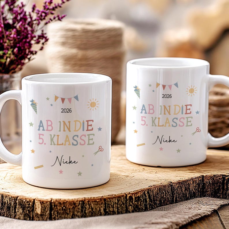 May include: Two white ceramic mugs with the text "AB IN DIE 5. KLASSE" in pastel colours, the year 2026 and the name "Nieke". The mugs are decorated with stars, carrots, scissors, and sun illustrations.