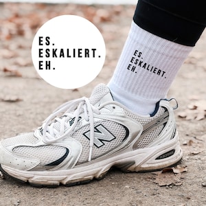 Könnte beinhalten: Weiße Sportsocken mit dem Text "ES. ESKALIERT. EH." werden mit einem weißen und silbernen Sportschuh getragen. Der Schuh hat weiße Schnürsenkel und ein großes "N"-Logo an der Seite. Der Schuh steht auf einem schmutzigen Untergrund.