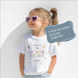 May include: A white t-shirt with colourful text and graphics, including the words "AB IN DIE 5. KLASSE" and the name "Nieke". The shirt has a festive design with stars, a sun, and a banner. The girl is wearing heart-shaped sunglasses.