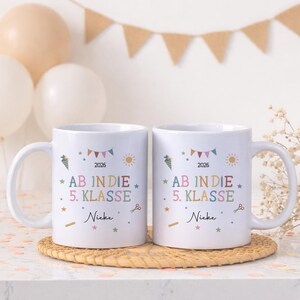 May include: Two white ceramic mugs with the text "AB IN DIE 5. KLASSE" and the name "Nieke" printed on them. The mugs have colorful illustrations of carrots, suns, stars, and scissors. The year 2026 is also printed on the mugs.
