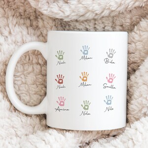 May include: White ceramic mug with colourful handprint designs and names such as Noah, Milan, and Bela. Each handprint has a heart in the centre. The mug has a large, curved handle.