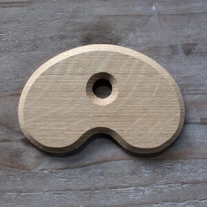May include: A light brown, kidney-shaped wooden piece with a smooth, rounded edge and a single circular hole centered near the top. The object features a visible wood grain texture and a beveled perimeter, presenting a flat, symmetrical form.