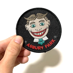 May include: A black embroidered patch with a cartoon face and the text "ASBURY PARK" in red.