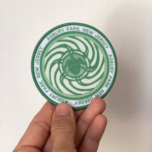 May include: A green and white circular patch with the text "ASBURY PARK, NEW JERSEY ASBURY PARK, NEW JERSEY" around the edge. The center of the patch features a swirling green design with a white face in the center.