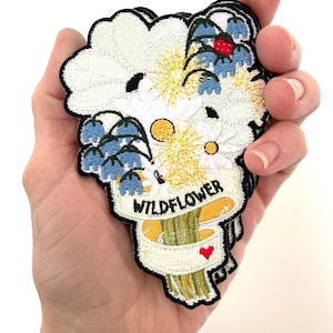 May include: A stack of embroidered iron-on patches featuring a bouquet of white, yellow, and blue flowers with a red heart and the text "WILD FLOWER".