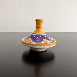 May include: A small yellow ceramic dish with a blue and red geometric pattern. The dish has a lid with a pointed top.