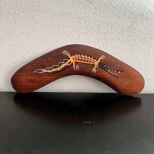 Puede incluir: Un boomerang de madera con un diseño pintado de un lagarto en rojo, amarillo, negro y blanco. El boomerang es marrón y tiene una forma curva.