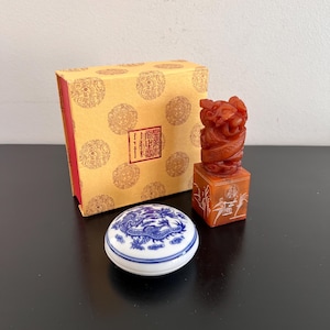 Puede incluir: Un conjunto de tres sellos de sello chino, uno en una caja de porcelana azul y blanca, otro en una caja roja y dorada, y otro tallado en piedra roja con un diseño de dragón. El sello de piedra roja está sobre una base con un diseño tallado de bambú.
