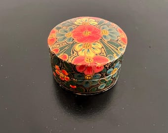 Pode incluir: Uma pequena caixa decorativa redonda com um design floral. A caixa é pintada com flores vermelhas, amarelas e azuis sobre um fundo verde escuro. A caixa está fechada e assenta numa superfície preta.