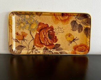 May include: A rectangular tray with a gold rim and a floral pattern. The pattern features brown, yellow, and white roses on a beige background.