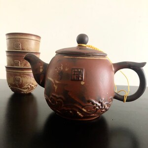 May include: A brown ceramic teapot with a wave design and a matching set of four teacups. The teapot has a lid with a small knob and a string attached to the lid. The teapot and cups are on a black surface.