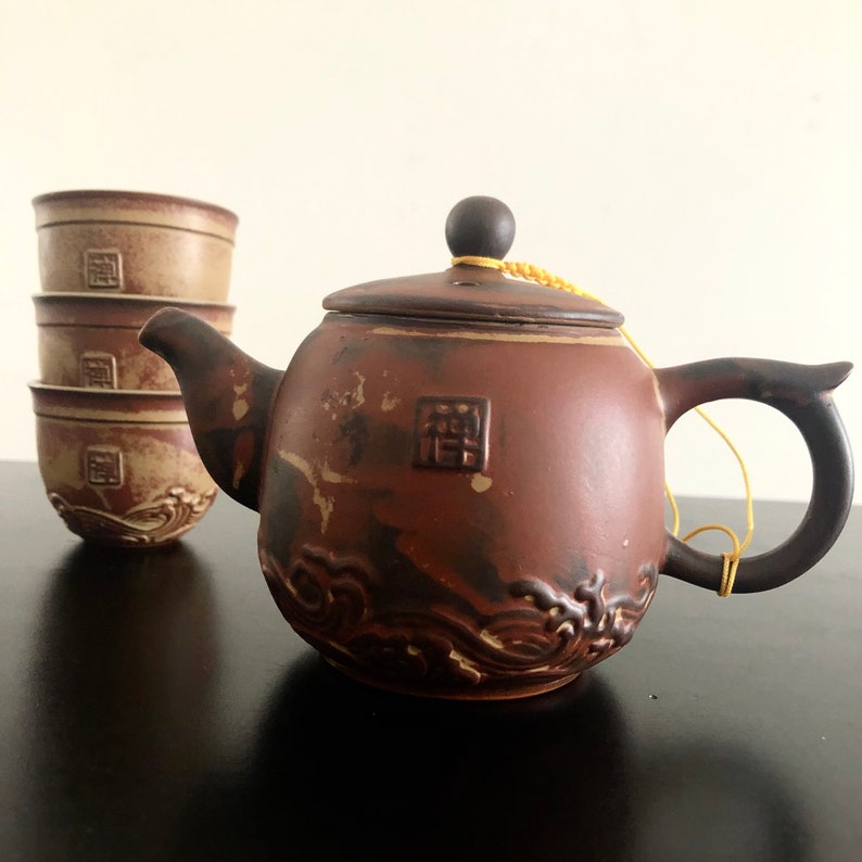 May include: A brown ceramic teapot with a wave design and a matching set of four teacups. The teapot has a lid with a small knob and a string attached to the lid. The teapot and cups are on a black surface.
