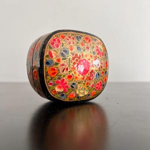 May include: A small, oval-shaped box with a black base and a colourful floral design. The box is decorated with intricate details in red, pink, yellow, green, blue, and gold.