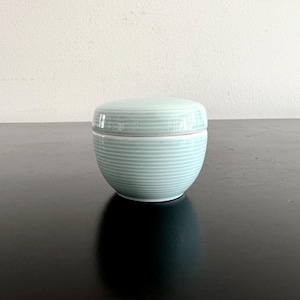 May include: A small, round, lidded ceramic container in a pale blue-green colour. The container and lid feature horizontal, evenly spaced, textured lines. The container sits on a dark, reflective surface.