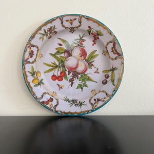 May include: A decorative plate with a white background and a colorful fruit design. The plate features peaches, cherries, and grapes, with a teal rim and gold accents. Butterflies and bees are also depicted in the design.