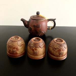 May include: A brown ceramic teapot with a matching set of four teacups. The teapot and cups have a wave design and a brown glaze.