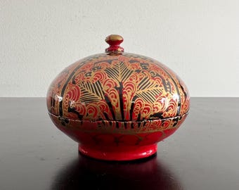 Pode incluir: Uma caixa decorativa redonda vermelha com tampa. A caixa é adornada com intrincados padrões dourados e pretos. A tampa tem um pequeno botão dourado. A caixa assenta numa base vermelha, criando um contraste marcante.