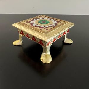 May include: A square gold-toned metal stand with four claw-shaped feet. The top surface features a decorative inlay with a central green circle surrounded by white and red floral-inspired patterns. The sides are accented with red enamel details.