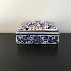 May include: A rectangular ceramic trinket box with a lid. The box is white with blue floral patterns and gold trim. The lid features a detailed floral design. The box is sitting on a black surface.
