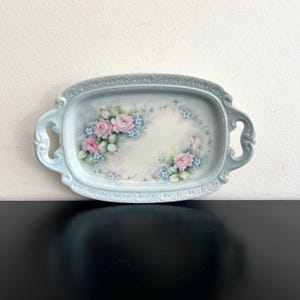 Puede incluir: Una bandeja de servicio ovalada azul claro con asas. La bandeja presenta un diseño floral de rosas rosas y nomeolvides azules. Los bordes de la bandeja están decorados con un delicado patrón en relieve.