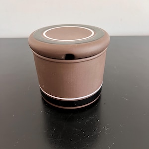 Könnte beinhalten: Eine braune Zuckerdose aus Keramik mit Deckel. Die Schale hat einen weißen und schwarzen Streifen um die Mitte und einen weißen Kreis mit einem grünen Ring um die Oberseite des Deckels.