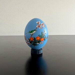 以下が含まれることがあります： 手描きの青い装飾用の卵。オレンジ色の花と白い花が咲く枝に、緑色の鳥がとまっています。卵は小さな濃い青色のスタンドに取り付けられており、自然にインスパイアされたデザインが施されています。