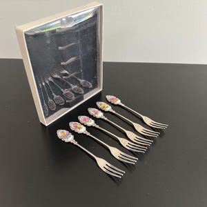 May include: Set of six small silver forks with floral designs on the handles. The forks are arranged on a black surface next to a box with a clear lid, which also contains forks. The floral designs include pink, yellow, and purple flowers.