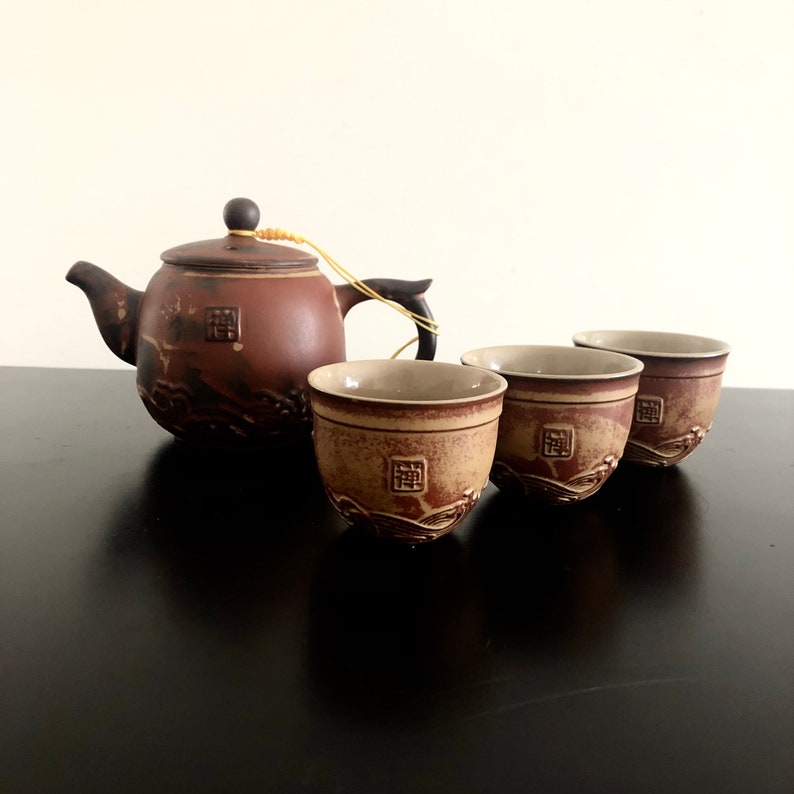 May include: A brown ceramic teapot with a matching set of three teacups. The teapot and cups have a decorative design of waves and a square symbol.