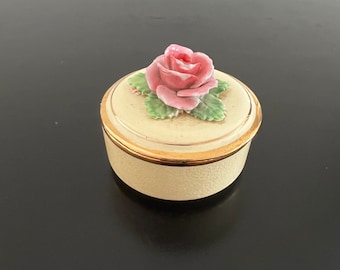 Puede incluir: Una pequeña caja redonda de color crema con un borde dorado y una rosa rosa en 3D en la tapa.