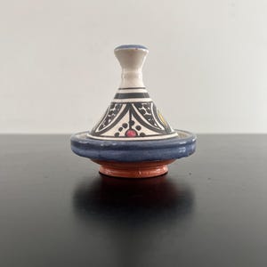 May include: A small, decorative ceramic tagine pot. The conical lid is white with black and red geometric patterns. The base is blue and brown. The tagine is a traditional cooking vessel, often used in Moroccan cuisine.
