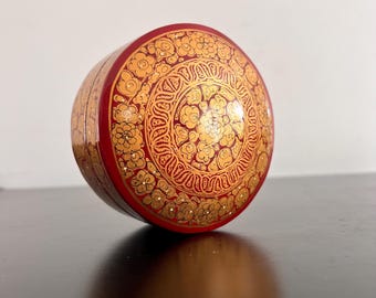 Pode incluir: Uma caixa redonda decorativa vermelha com um design floral dourado. A caixa é feita de papel maché e tem um acabamento brilhante.