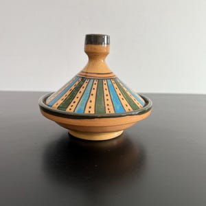 May include: A ceramic tagine with a conical lid featuring alternating vertical stripes in shades of blue, green, and tan. The lid is topped with a dark brown knob, and the base has a matching dark brown rim, showcasing a hand-painted dotted pattern.