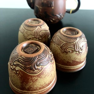 May include: Three brown ceramic teacups with a wave pattern and a small square design on the top. The cups are on a black surface.