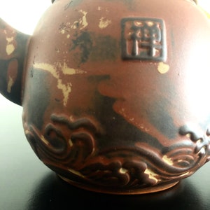 May include: A brown ceramic teapot with a wave design and a square seal with the character "禪" in black.