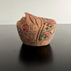 May include: A carved wooden box with a lid, featuring a face design. The box is primarily brown with intricate floral and geometric patterns in red, green, and tan. The lid is shaped to resemble a profile.