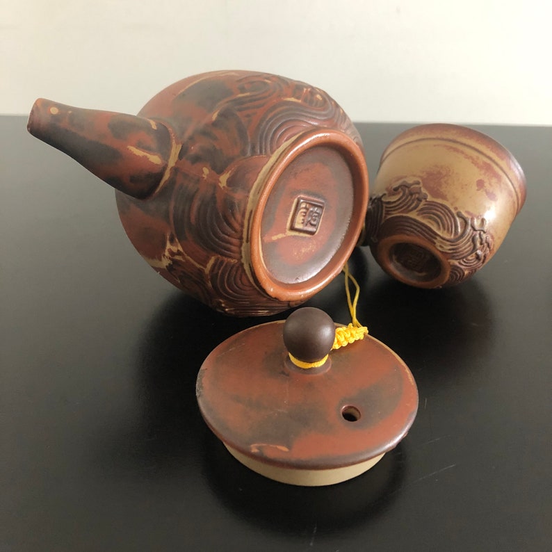 May include: A brown ceramic teapot with a lid and a matching cup. The teapot has a decorative design of waves and the cup has a similar design. The teapot has a small handle and a spout. The lid has a knob on top. The teapot and cup are connected by a yellow string.
