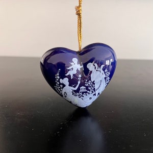 May include: A blue heart-shaped ornament with a white design of two figures and a cupid. The ornament has a gold string for hanging.