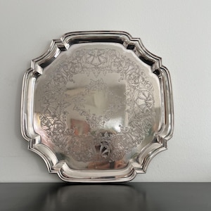May include: A silver serving tray with a decorative, scalloped edge. The tray has an engraved floral and scroll pattern in the centre. The tray is square with rounded corners.