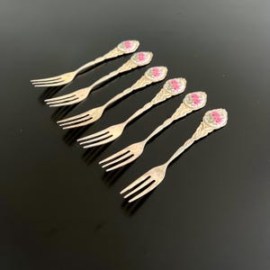 May include: Six antique silver forks with floral designs on the handles. Each fork has a three-tine design and a decorative handle with a pink and white floral pattern. The forks are arranged on a black surface.