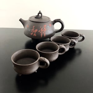 May include: A brown ceramic teapot with a lid and a handle, along with four matching teacups. The teapot and cups have a decorative design of red and green floral patterns.