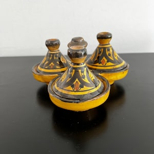 May include: Four small yellow and black ceramic tagines with intricate designs. The tagines are arranged in a circle on a black surface.
