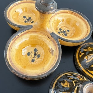 May include: A ceramic serving dish with three individual bowls and a central handle. The bowls are yellow with black floral designs. The handle is black and brown.