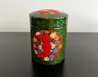 Pode incluir: Uma caixa cilíndrica verde com um desenho floral pintado no lado. As flores são vermelhas, amarelas, azuis e brancas. A caixa tem uma tampa com uma borda dourada.