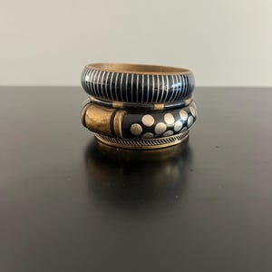 May include: A stack of three bangles featuring a top bracelet with black and silver vertical stripes, a middle bracelet with black and silver polka dots and a gold-toned accent, and a bottom bracelet with a textured gold-toned metal finish.