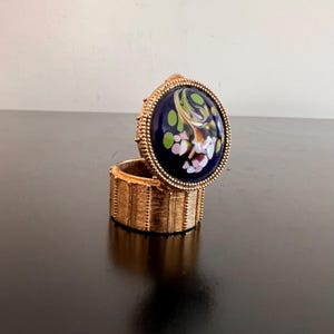 May include: A small, round, gold-coloured trinket box with a hinged lid. The lid is decorated with a blue enamel background and a colourful floral design.